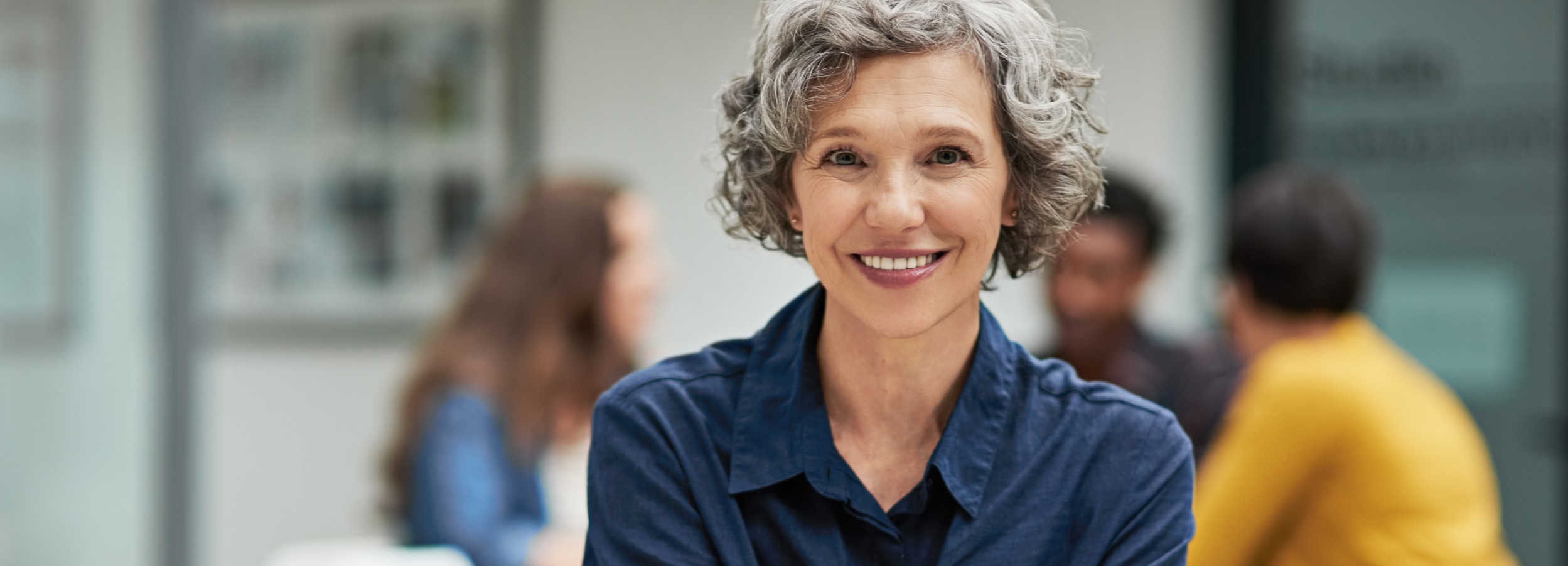 Das Bild zeigt eine lächelnde, selbstbewusste Frau in einer Büroumgebung, während im Hintergrund ein unklares Teammeeting stattfindet.

Im Vordergrund des Bildes ist eine Frau mittleren Alters mit kurzem, gewelltem, grauem Haar zu sehen. Sie trägt ein blaues Hemd und lächelt freundlich in die Kamera. Ihr Lächeln wirkt natürlich und einnehmend. Ihre Augen sind wach und aufmerksam. Im Hintergrund sind verschwommen weitere Personen zu erkennen, die vermutlich an einem Meeting teilnehmen. Eine Person trägt ein gelbes Oberteil, eine andere ein blaues. Der Hintergrund ist hell und modern gestaltet, mit unscharfen Bildern oder Grafiken an der Wand. Die Atmosphäre wirkt professionell, aber gleichzeitig entspannt und positiv. Die Stimmung ist einladend und kompetent, was vor allem durch das Lächeln der Frau im Vordergrund verstärkt wird.
