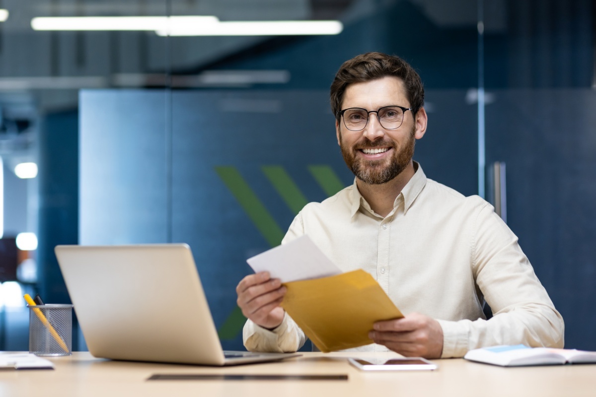 Junger Mann sitzt glücklich an seinem Schreibtisch im Büro und freut sich über den Inhalt eines geöffneten Briefs, den er in der Hand hält. Ein geöffneter Laptop steht vor ihm.