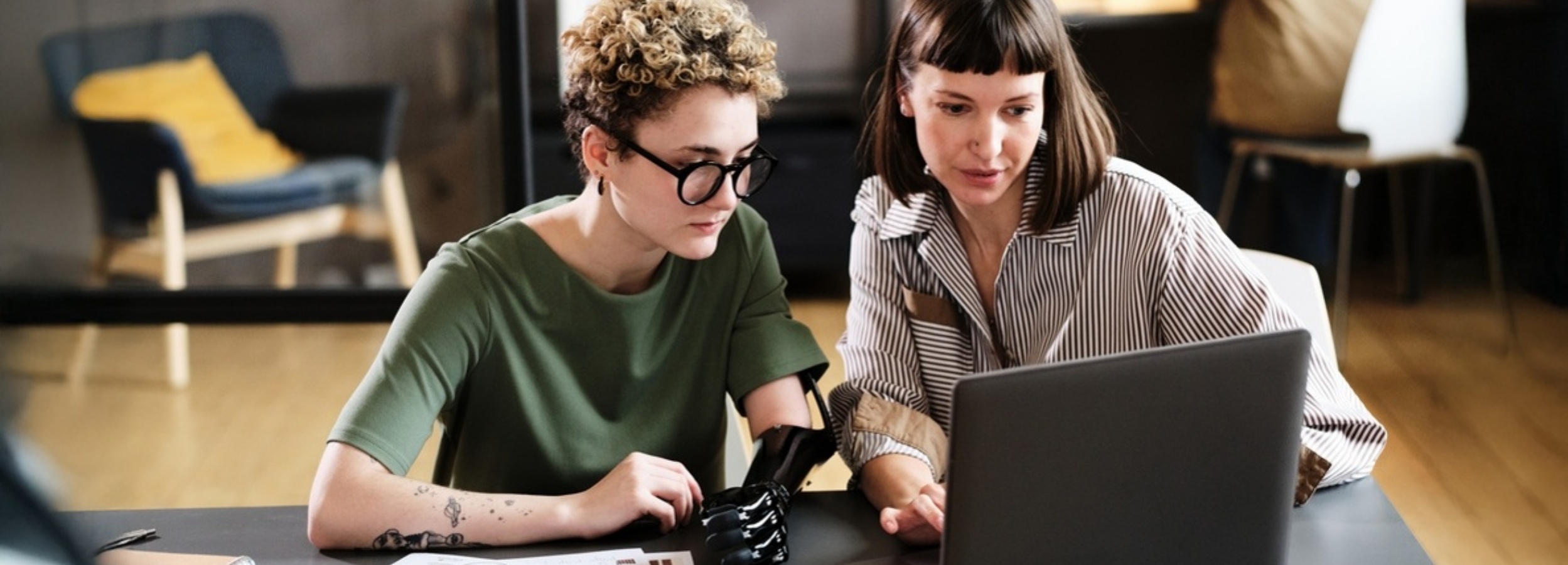 Zwei Frauen, von denen eine eine Armprothese trägt, arbeiten konzentriert an einem Laptop in einer modernen Büroumgebung zusammen.

Auf dem Bild sind zwei Frauen zu sehen, die an einem dunklen Tisch sitzen und auf den Bildschirm eines geöffneten, dunkelgrauen oder schwarzen Laptops blicken. Die Frau auf der linken Seite ist jung, hat kurze, lockige, hellblonde Haare und trägt eine runde, dunkle Brille. Sie ist in einem olivgrünen, kurzärmeligen Oberteil gekleidet. Ihr linker Unterarm ist eine hochmoderne, glänzende, schwarze Armprothese, die detailreich gestaltet ist und die Form einer Hand andeutet. Auf ihrem rechten Unterarm sind mehrere kleine Tätowierungen zu erkennen, die wie kleine Figuren oder Symbole aussehen. Ihr Blick ist fokussiert auf den Laptop gerichtet.

Neben ihr, auf der rechten Seite des Bildes, sitzt eine zweite Frau mittleren Alters mit glattem, dunklem Haar und einem geraden Pony. Sie trägt ein Hemd mit feinen, dunklen und hellen Längsstreifen. Ihre rechte Hand ist leicht angehoben, und ihr Zeigefinger scheint auf den Laptop-Bildschirm zu deuten oder etwas darauf zu zeigen. Auch ihr Gesichtsausdruck ist konzentriert und engagiert, während sie auf den Bildschirm schaut.

Im leicht unscharfen Hintergrund links ist ein bequemer, blauer Sessel mit einem leuchtend gelben Kissen zu erkennen, dessen Holzfüße sichtbar sind. Im rechten Hintergrund sind weitere Möbelstücke angedeutet, darunter ein Stuhl mit einer hellen Sitzfläche. Der Boden scheint aus hellem Holz zu sein. Die Beleuchtung ist hell und natürlich, was auf eine moderne Büro- oder Co-Working-Umgebung hindeutet.

Die Atmosphäre des Bildes ist professionell und kollaborativ. Es strahlt eine Stimmung der Konzentration, des Engagements und der effektiven Zusammenarbeit aus, wobei der Fokus auf der gemeinsamen Aufgabe und der Nutzung von Technologie liegt. Die Darstellung der Frau mit der Prothese unterstreicht zudem ein Thema der Inklusion und modernen Arbeitswelt.