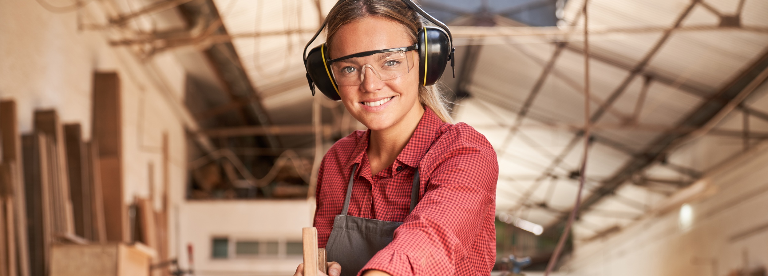Eine junge Frau strahlt zuversichtlich in die Kamera, während sie in ihrer Werkstatt arbeitet.

Das Bild zeigt eine junge Frau, die in einer Schreinerei oder Holzarbeitswerkstatt steht und in die Kamera lächelt. Sie trägt eine rote karierte Bluse, eine graue Schürze, eine Schutzbrille und ein Paar schwarze Gehörschutzkopfhörer, die über ihren blonden Zopf reichen. Ihre Kleidung und die Schutzausrüstung deuten darauf hin, dass sie mit Holz arbeitet oder andere handwerkliche Tätigkeiten ausübt, die Gehör- und Augenschutz erfordern. Ihre Haltung ist aufrecht und sie hält ein Stück Holz in ihren Händen, was darauf hindeutet, dass sie gerade bei der Arbeit ist oder diese vorbereitet.

Hinter ihr ist eine unscharfe Kulisse einer Werkstatt zu sehen, die mit Holzbrettern und anderen Materialien gefüllt ist, die vertikal an den Wänden lehnen oder in Stapeln gelagert sind. Die Decke der Werkstatt ist im Hintergrund sichtbar und zeigt eine industriell anmutende Konstruktion aus Metallträgern und Dachplatten.

Die Atmosphäre des Bildes ist positiv und ermutigend. Die Frau lächelt offen und freundlich, was eine einladende und selbstbewusste Ausstrahlung vermittelt. Die Beleuchtung ist hell und natürlich, was die Szene warm und einladend erscheinen lässt. Die unscharfen Hintergründe lenken den Fokus auf die Frau und ihre Arbeit und verleihen dem Bild eine professionelle Tiefenwirkung.

Insgesamt vermittelt das Bild eine Botschaft von Kompetenz, Selbstständigkeit und der Freude an manueller Arbeit, möglicherweise auch eine feministische Aussage, die Frauen in traditionell männlich dominierten Berufen zeigt.