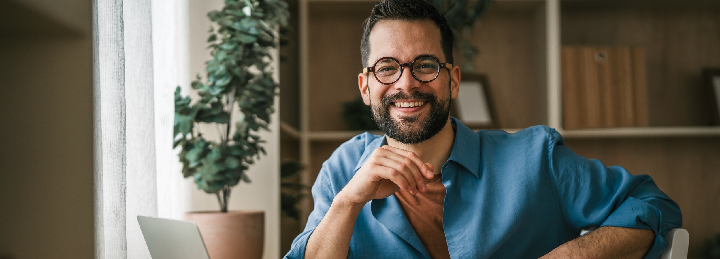 Ein Mann mit Brille und Bart lächelt in die Kamera, während er an seinem Laptop arbeitet, was eine positive und produktive Arbeitsatmosphäre im Homeoffice signalisiert.

Das Bild zeigt einen Mann mittleren Alters, der einen blauen Hemd über einem orangefarbenen T-Shirt trägt. Er hat einen gepflegten Bart und trägt runde, braune Brillengläser. Seine Arme sind vor ihm verschränkt, und er sitzt an einem Tisch, auf dem ein silberner Laptop geöffnet ist. Er blickt direkt in die Kamera und lächelt warmherzig, was eine freundliche und zugängliche Ausstrahlung vermittelt.

Im Hintergrund ist ein Fenster mit hellem Licht zu sehen, das eine sanfte Beleuchtung für die Szene liefert. Daneben steht eine Topfpflanze mit üppig grünen Blättern, die dem Raum Leben und Frische verleiht. Weiter im Hintergrund sind Regale mit Ordnern und Dekorationen zu erkennen, was auf eine Büroumgebung oder ein Arbeitszimmer hindeutet. Eine weiße Stuhllehne ist am rechten Bildrand sichtbar.

Die Atmosphäre des Bildes ist entspannt, positiv und produktiv. Das Lächeln des Mannes und die helle, natürliche Beleuchtung schaffen eine einladende und angenehme Stimmung, die typisch für ein gut eingerichtetes Homeoffice ist. Es vermittelt ein Gefühl von Zufriedenheit und Engagement bei der Arbeit.