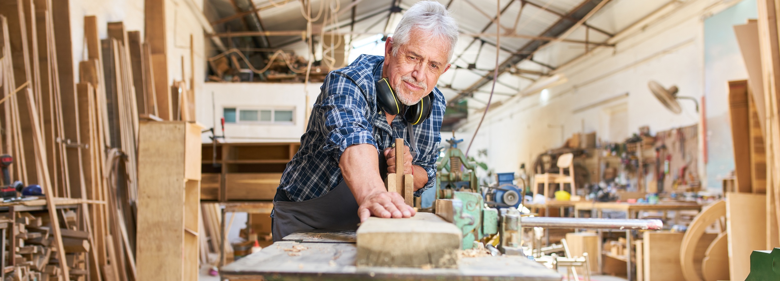 Ein älterer Handwerker arbeitet konzentriert in seiner Werkstatt an einem feinen Holzstück.

Das Bild zeigt einen älteren Mann mit grauem Haar und einem karierten Hemd, der in einer Werkstatt steht und ein Holzstück bearbeitet. Er trägt Kopfhörer um den Hals. Seine Hände sind auf dem Holzstück und einem Stechbeitel platziert. Im Hintergrund sind Regale voller Holzstücke und Werkzeuge zu sehen. Die Werkstatt wirkt belebt und voller Arbeitsmaterialien. Die Atmosphäre ist handwerklich und professionell, geprägt von der Hingabe des Mannes zu seinem Handwerk.