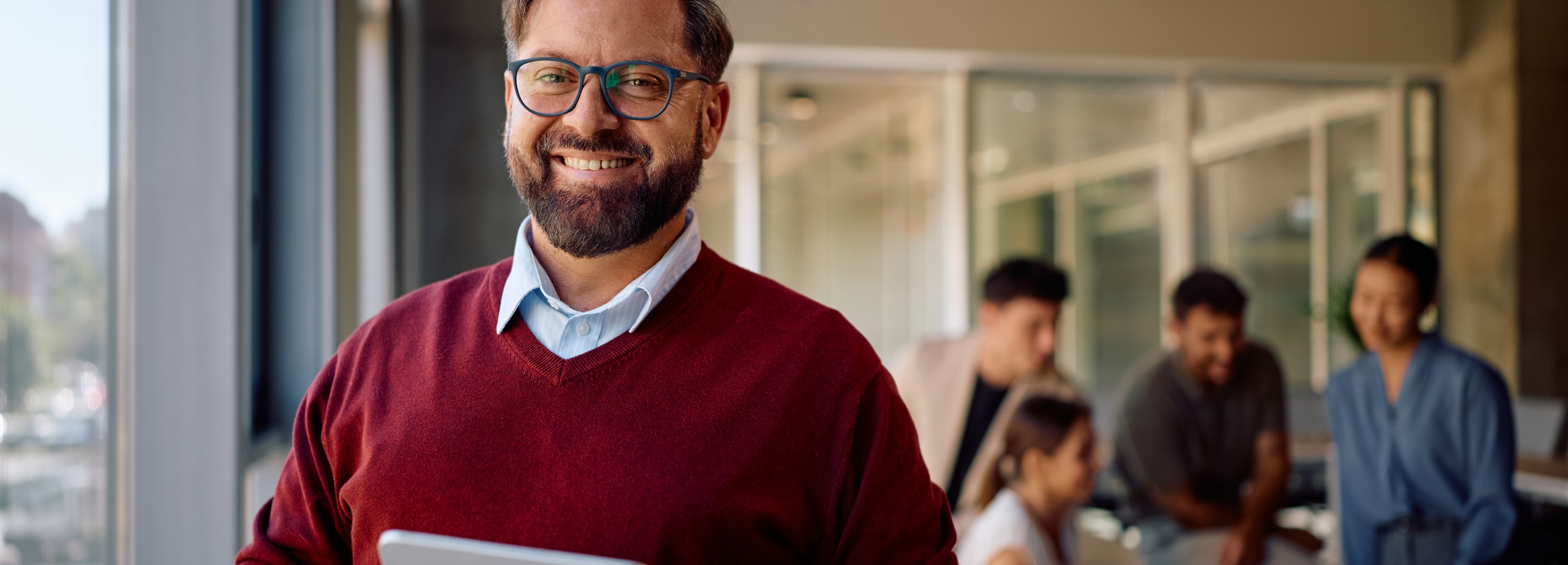 Ein Mann mit Brille lächelt in die Kamera, während er im Hintergrund ein unscharfes Teammeeting leitet.

Das Bild zeigt einen Mann mit Bart und Brille, der freundlich lächelt und ein Tablet hält, während im Hintergrund eine Gruppe von vier Personen in einem Meetingraum zu sehen ist. Der Mann trägt einen roten Pullover über einem hellblauen Hemd. Im Hintergrund sitzen oder stehen die Teammitglieder und scheinen in ein Gespräch vertieft zu sein. Die Atmosphäre wirkt professionell und entspannt zugleich, mit einem Fokus auf Zusammenarbeit und positiver Kommunikation. Das Licht ist hell und natürlich, was die offene und einladende Stimmung des Raumes unterstreicht.