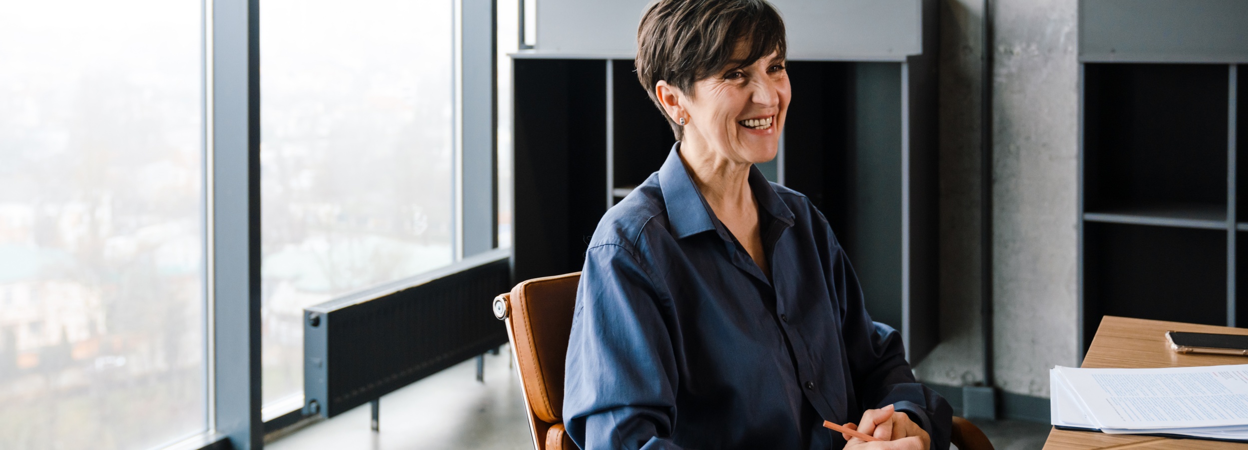 Das Bild zeigt eine lächelnde, reife Frau, die in einem modernen Büro sitzt.

Im Detail zeigt das Bild eine Frau mittleren Alters mit kurzen, dunklen Haaren, die ein dunkles, knielanges Hemd trägt, während sie auf einem braunen Lederstuhl an einem Holztisch sitzt. Ihr Gesicht strahlt ein offenes Lächeln aus, und sie scheint in ein Gespräch vertieft zu sein. Auf dem Tisch vor ihr liegen gestapelte Dokumente, ein Smartphone und ein brauner Bleistift, den sie in ihren Händen hält. Im Hintergrund befindet sich ein großes Fenster, das einen verschwommenen Blick auf eine Stadtlandschaft bietet, sowie ein modernes Regal mit dunklen Fächern. Die Atmosphäre ist hell und professionell, mit einem Hauch von Wärme und Vertrautheit durch das Lächeln der Frau. Die Stimmung wirkt positiv und einladend, was auf ein angenehmes Arbeitsumfeld oder ein entspanntes Gespräch hindeutet.