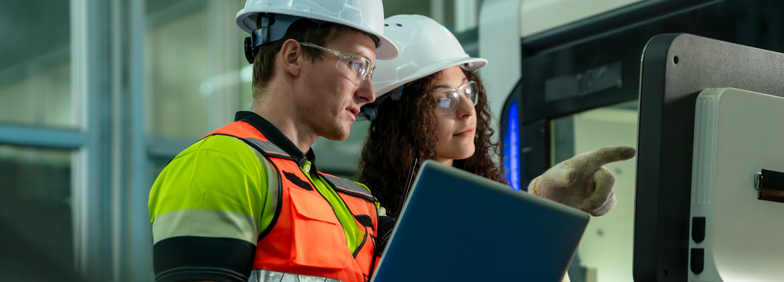 Zwei Ingenieure, ausgestattet mit Sicherheitsausrüstung, arbeiten an einer hochmodernen Maschine, wobei einer auf den Bildschirm deutet und der andere einen Laptop hält.

Das Bild zeigt zwei Personen, vermutlich Ingenieure oder Techniker, die eng zusammenarbeiten. Beide tragen weiße Schutzhelme und Schutzbrillen. Der Mann auf der linken Seite trägt eine auffällige Warnweste in Gelb und Orange über seinem Hemd. Er hat einen Laptop in der Hand. Die Frau auf der rechten Seite hat lockiges Haar und trägt einen Arbeitshandschuh an der Hand, mit der sie auf einen Bildschirm oder eine Maschine deutet.

Hinter den beiden Personen ist ein Teil einer großen, modernen Maschine sichtbar. Der Hintergrund ist unscharf und deutet auf eine industrielle Umgebung hin, möglicherweise eine Fabrik oder Werkshalle. Die Beleuchtung ist hell und gleichmäßig, was eine professionelle und konzentrierte Atmosphäre schafft. Die Stimmung ist arbeitsorientiert und kooperativ, da die beiden Personen in die Arbeit an der Maschine vertieft sind.